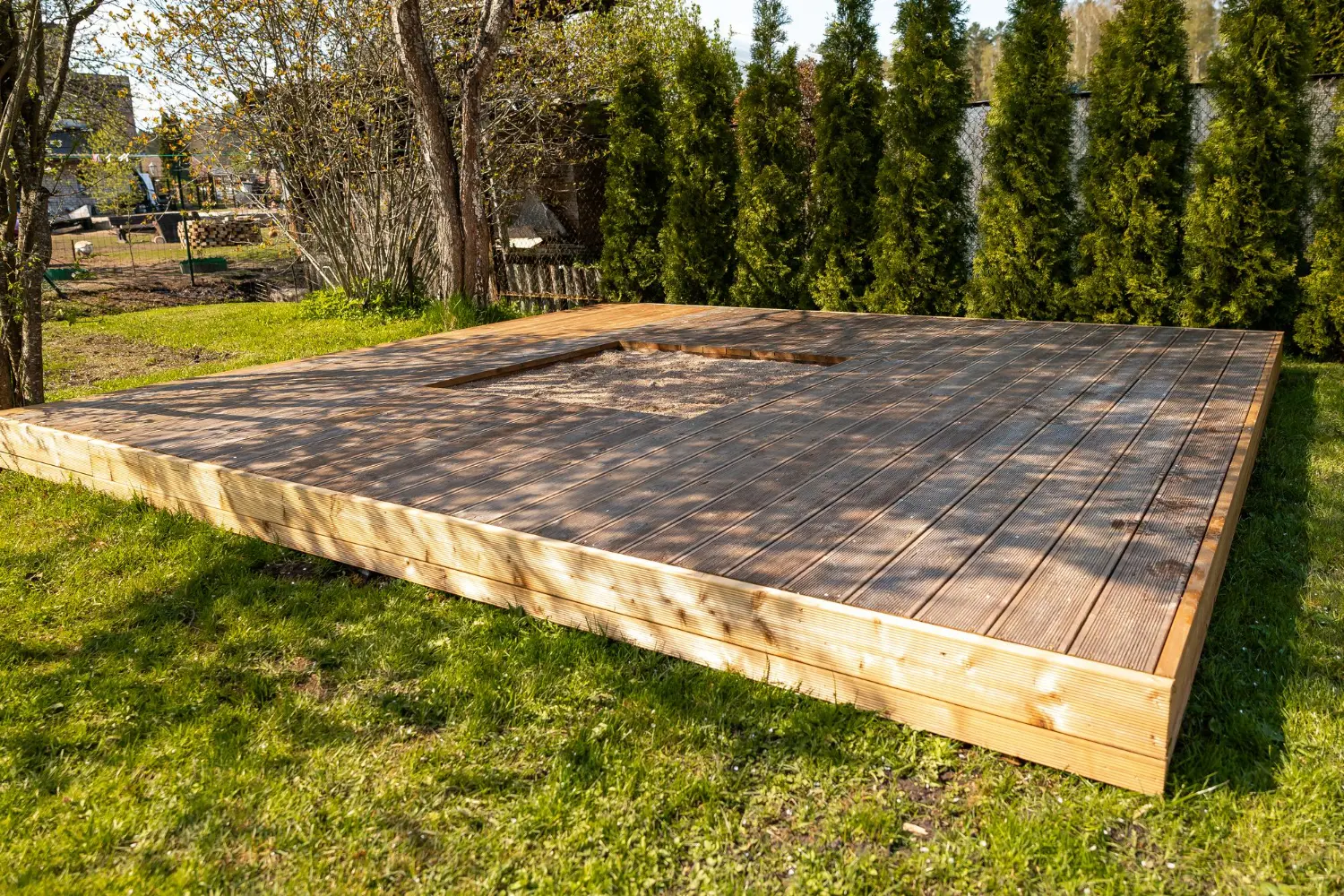 Drewniana platforma z kwadratowym wycięciem pośrodku, otoczona trawą i drzewami. tarasy szczecin