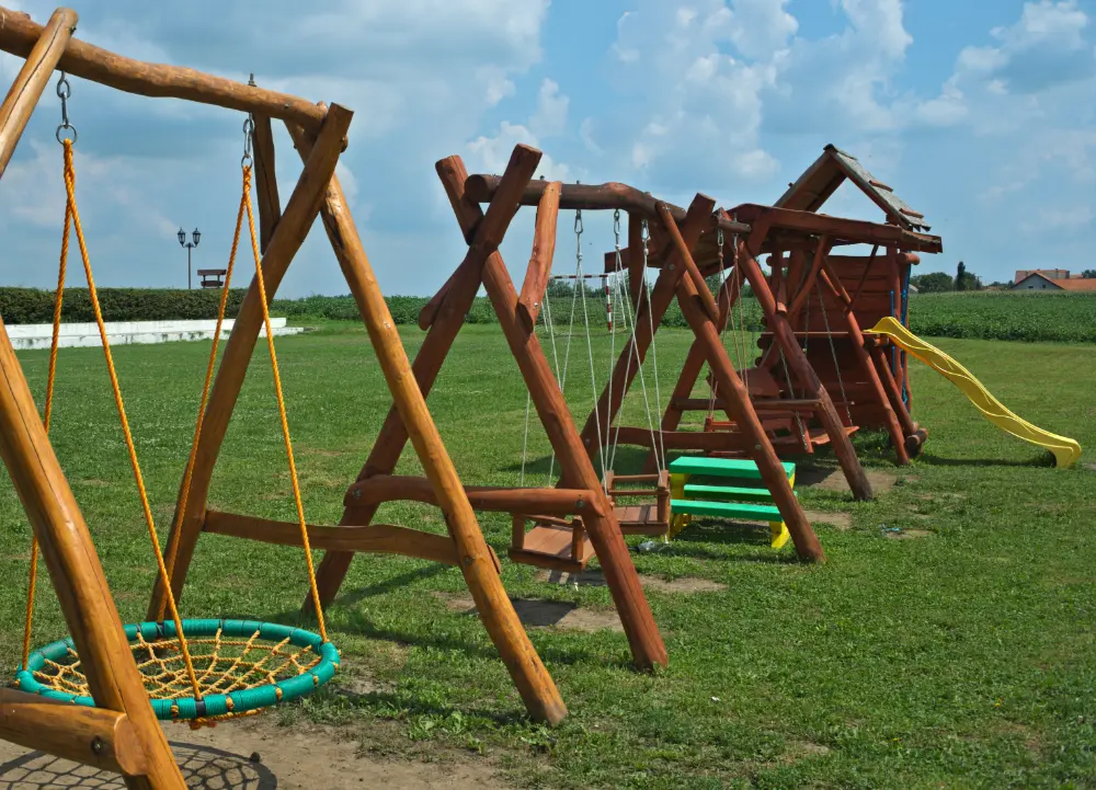 Drewniany plac zabaw z huśtawkami, zjeżdżalnią i ławkami na trawiastym boisku pod częściowo zachmurzonym niebem. tarasy szczecin