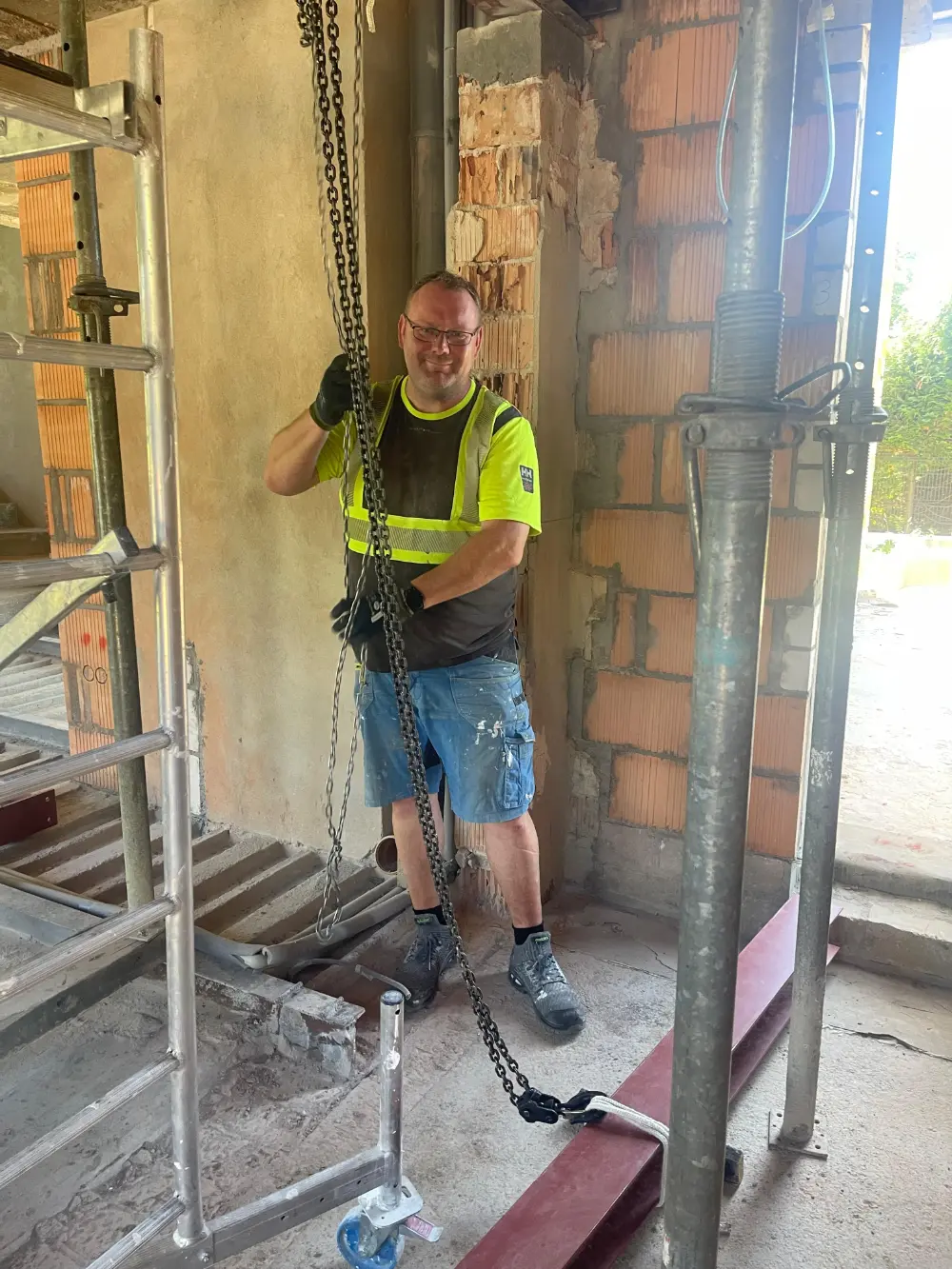 Uśmiechnięty pracownik budowlany w sprzęcie ochronnym trzymający łańcuch wewnątrz częściowo zbudowanej konstrukcji. tarasy szczecin