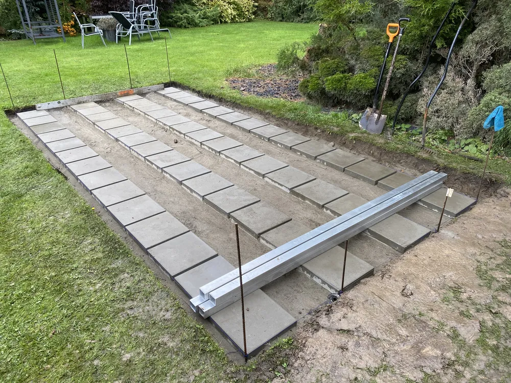 Prostokątne patio w budowie z kwadratowymi betonowymi kostkami brukowymi, metalowymi belkami, łopatami i trawą wokół. tarasy szczecin